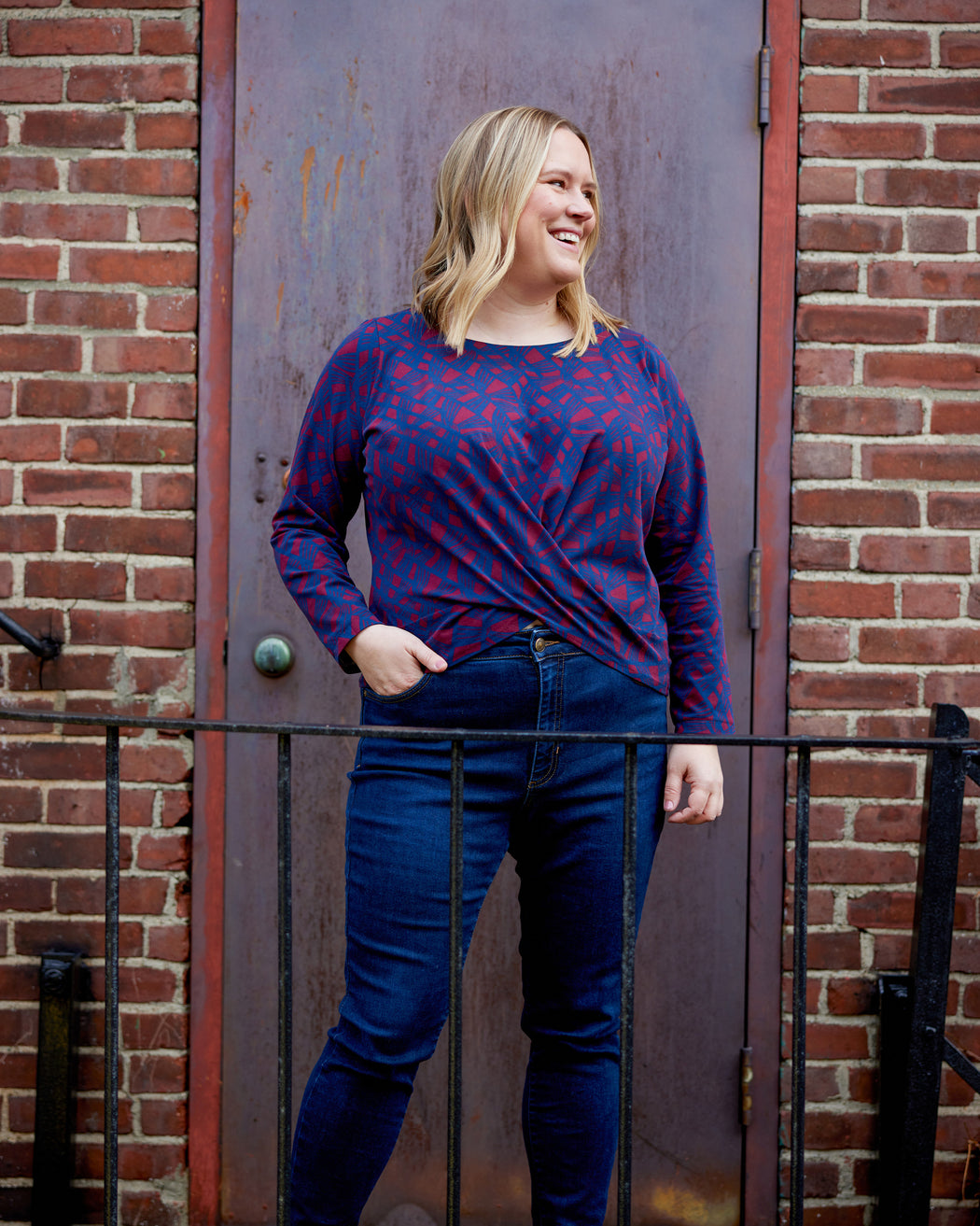 Woman wearing a red and blue patterned long-sleeve shirt and blue jeans standing against a brick wall. This top is the Brattle Twist Top sewing pattern from Cashmerette.