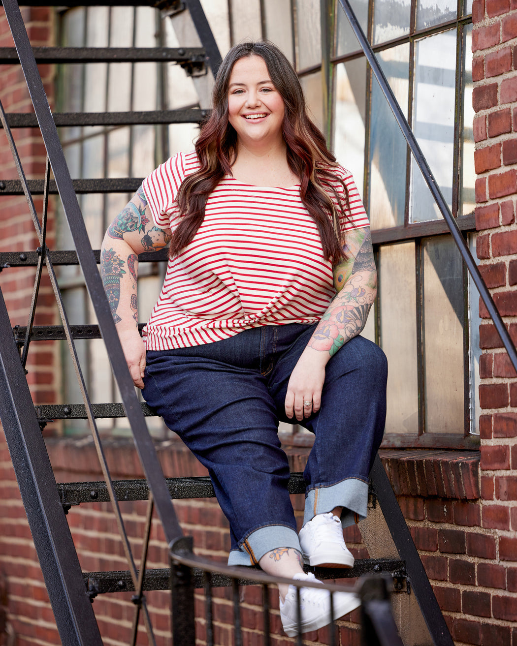 Woman in a red and white striped shirt and blue jeans sitting on metal stairs against a brick wall. This top is the Brattle Twist Top sewing pattern from Cashmerette.