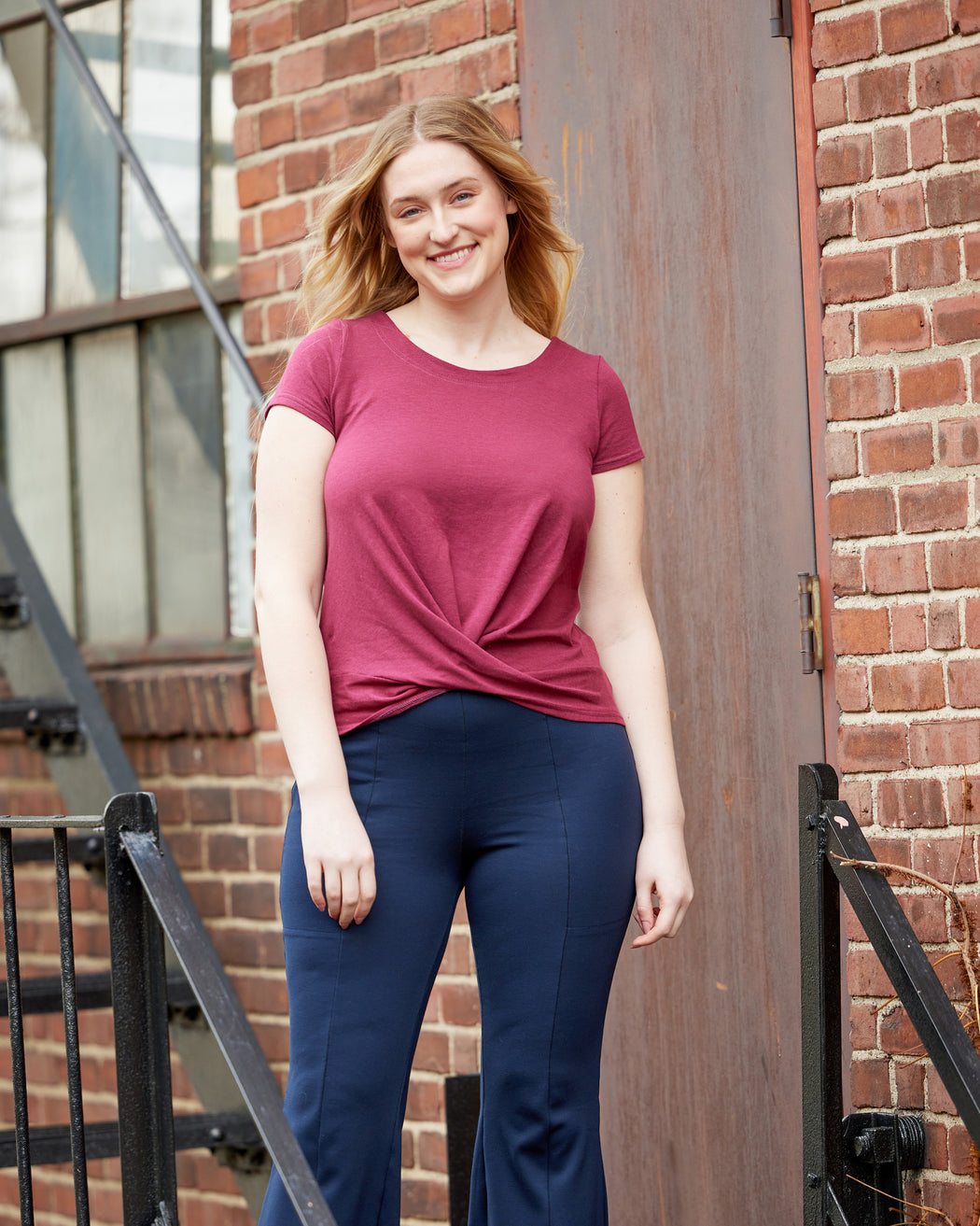 Woman wearing a red shirt and blue pants standing against a brick wall. This top is the Brattle Twist Top sewing pattern from Cashmerette.