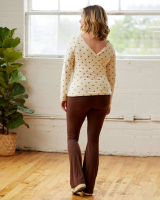 Woman wearing a patterned top and brown pants standing in a room with a large window and plant. This top is the Kenwin Raglan T-Shirt sewing pattern from Cashmerette.