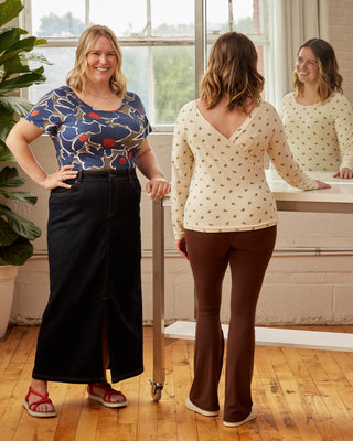 Two women standing in a room with large windows, one wearing a floral top and black pants, the other in a white patterned top and brown pants. This top is the Kenwin Raglan T-Shirt sewing pattern from Cashmerette.