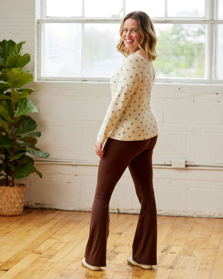 Woman wearing a white blouse with black patterns and brown pants standing in a room with a large window and plant. This top is the Kenwin Raglan T-Shirt sewing pattern from Cashmerette.