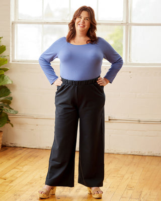 Woman wearing a blue top and black pants standing in a room with large windows. This top is the Kenwin Raglan T-Shirt sewing pattern from Cashmerette.