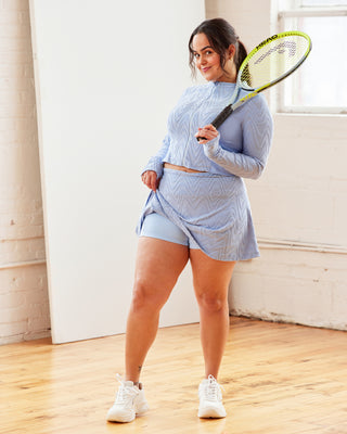 Woman in light blue athletic outfit holding a racquet in a room with white walls and wooden floor.  This top is the Bonair Athletic Top sewing pattern from Cashmerette.