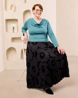 An older woman with short auburn hair and glasses is posing in a studio setting, wearing a teal cowl-neck long-sleeve top and a long black pleated skirt with a dark gray floral pattern. She is standing next to a white chair against a beige wall with sculptural recesses.  This is the Amesbury Cowl Top sewing pattern from Cashmerette.