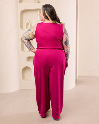 A woman with tattooed arms is shown from behind wearing a bright fuchsia sleeveless jumpsuit with wide-leg pants, standing in a minimalist beige room. This is the Amesbury Cowl Top sewing pattern from Cashmerette.
