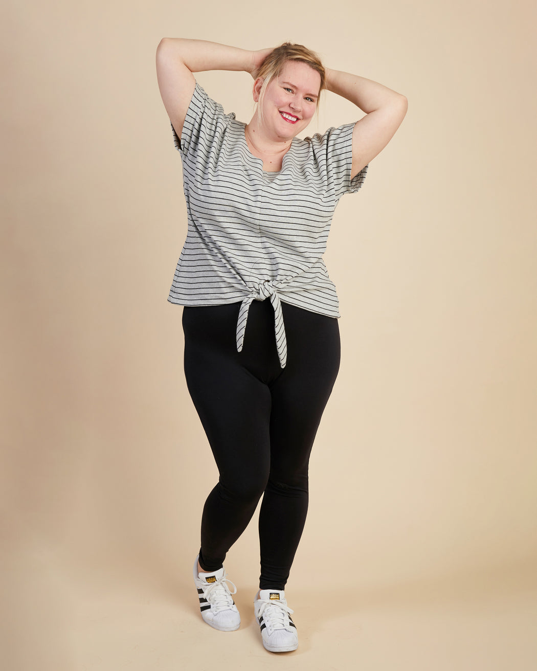 Woman wearing a striped shirt and black leggings on a beige background. This top is the Cedar Dolman Top sewing pattern from Cashmerette.