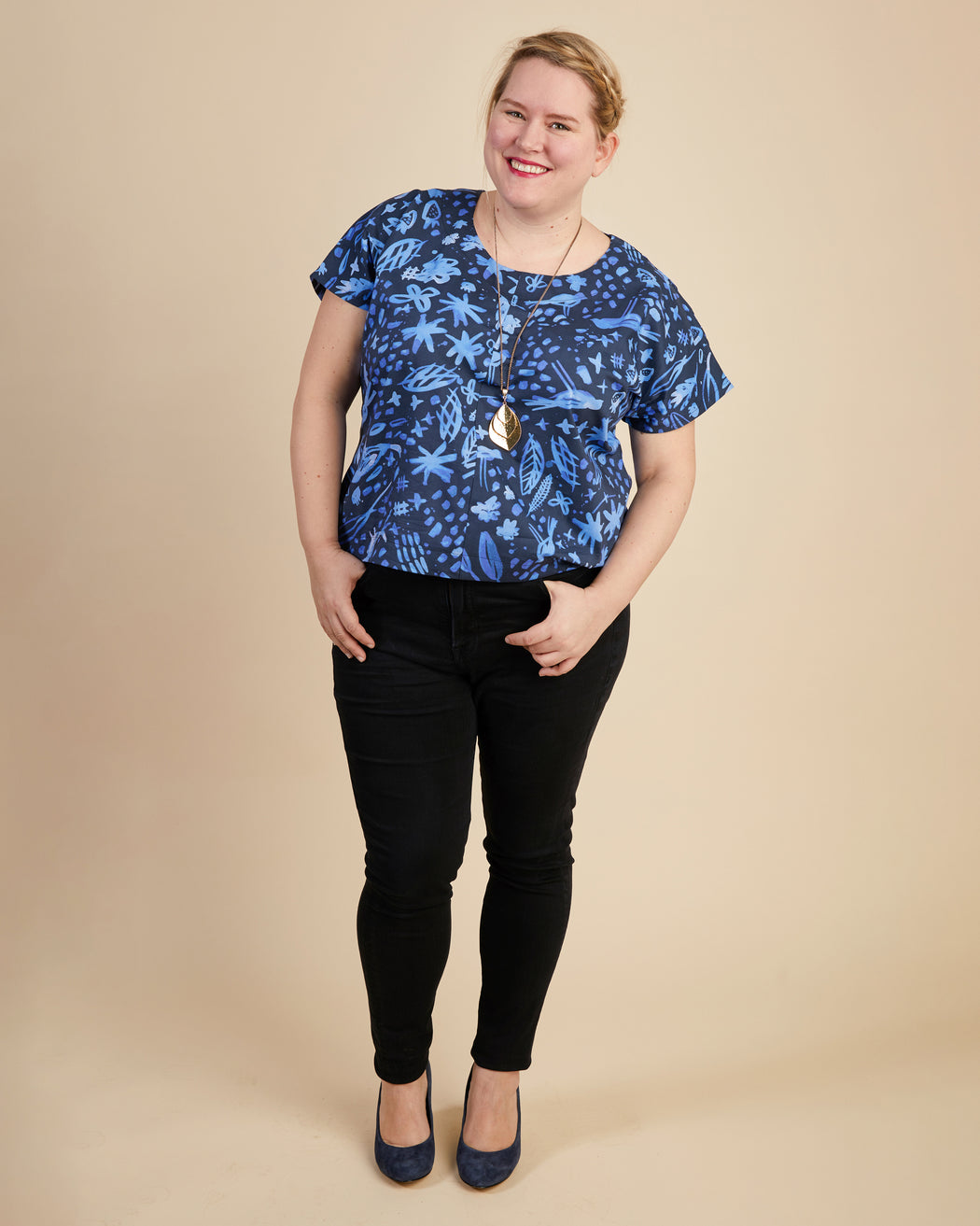 Woman wearing a blue floral blouse and black pants on a beige background. This top is the Cedar Dolman Top sewing pattern from Cashmerette.