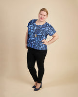 Woman wearing a blue floral top and black pants on a beige background. This top is the Cedar Dolman Top sewing pattern from Cashmerette.