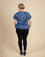 Person wearing a blue patterned top and black pants on a beige background. This top is the Cedar Dolman Top sewing pattern from Cashmerette.