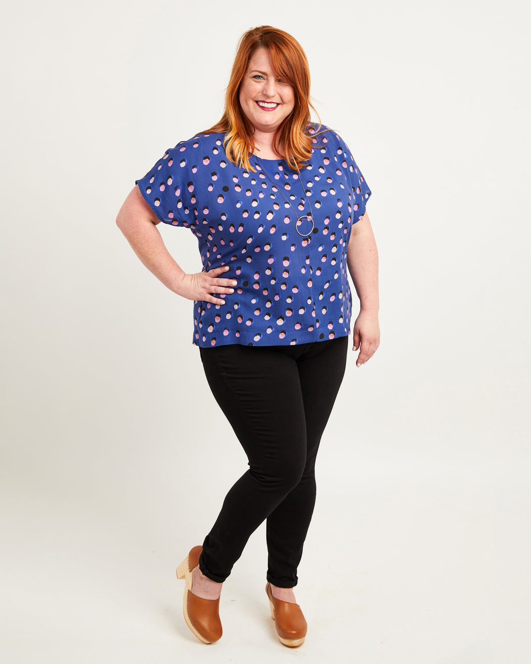 Woman wearing a blue polka dot top and black leggings on a white background. This top is the Cedar Dolman Top sewing pattern from Cashmerette.