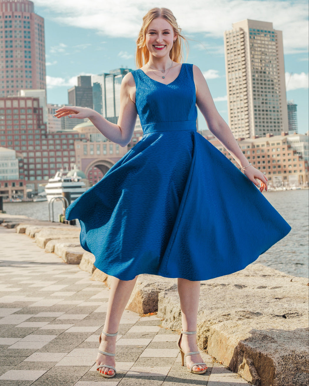 Woman in a blue dress standing on a waterfront with city skyline in the background. This skirt is the Gilmore Circle Skirt sewing pattern from Cashmerette.