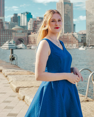 Woman in a blue dress standing by a waterfront with city skyline in the background. This skirt is the Gilmore Circle Skirt sewing pattern from Cashmerette.
