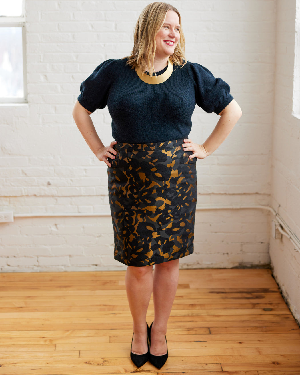 Woman wearing a navy top and leopard print skirt in a room with wooden floor and white brick wall. This skirt is the Elmwood Pencil Skirt sewing pattern from Cashmerette.
