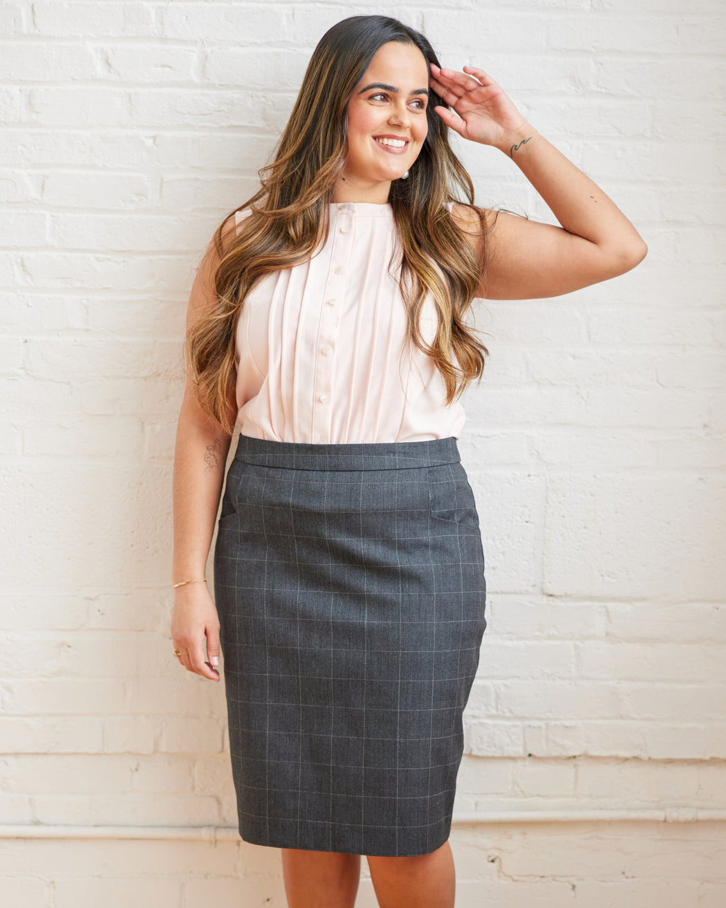 Woman wearing a white blouse and gray skirt against a white brick wall. This skirt is the Elmwood Pencil Skirt sewing pattern from Cashmerette.