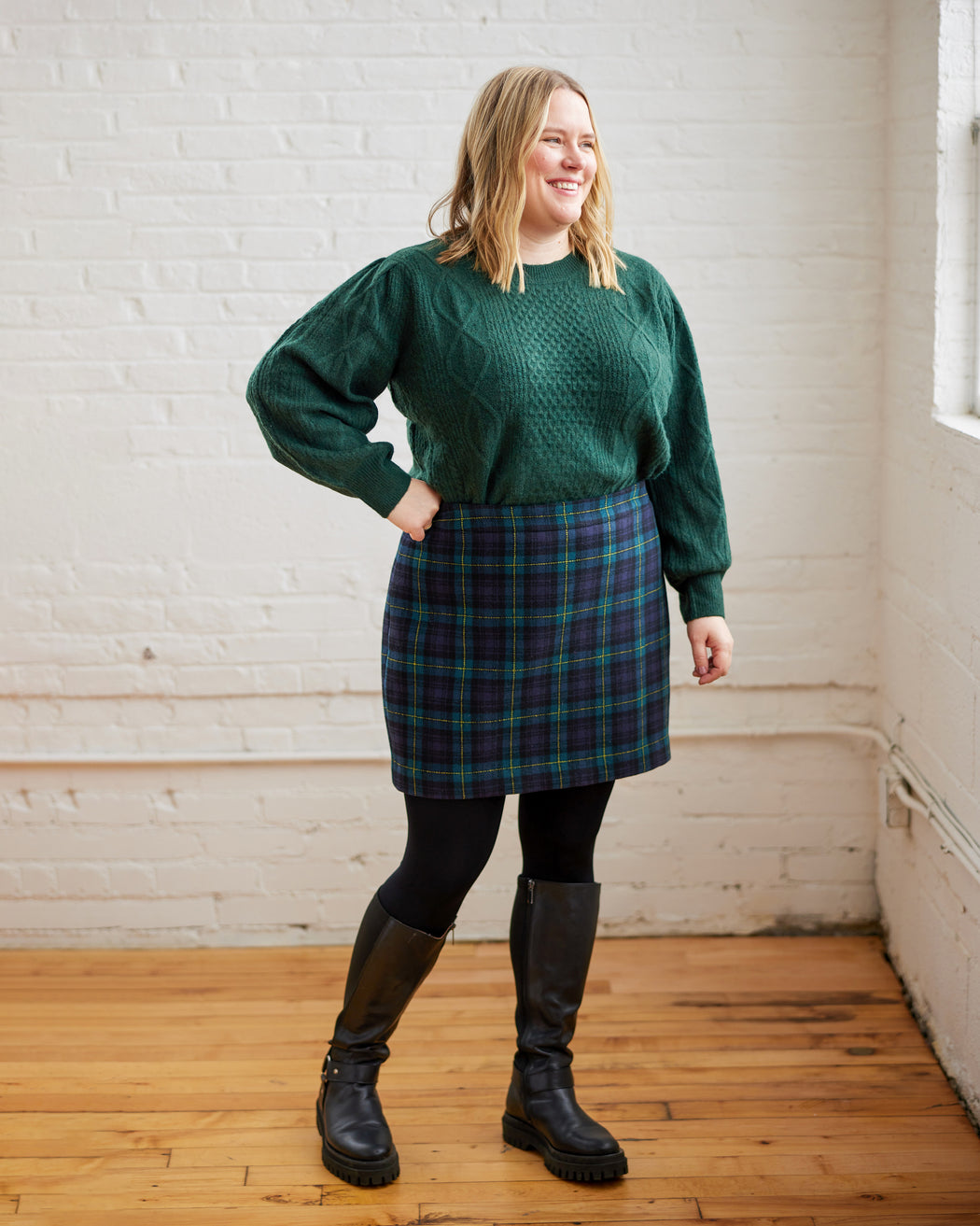 Person wearing a green sweater and plaid skirt standing in a room with white brick walls and wooden floors. This skirt is the Elmwood Pencil Skirt sewing pattern from Cashmerette.