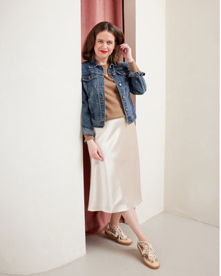 Woman wearing a denim jacket, brown top, white skirt, and sandals standing in a doorway. This skirt is the Astell Bias Skirt sewing pattern from Cashmerette.