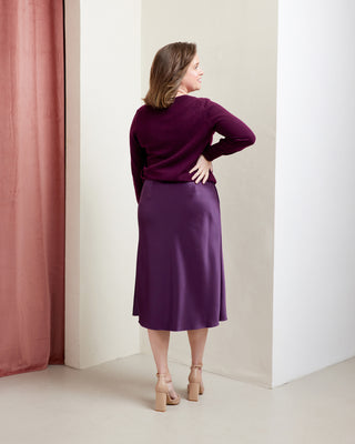 Woman wearing a purple outfit standing against a white wall with a pink curtain. This skirt is the Astell Bias Skirt sewing pattern from Cashmerette.