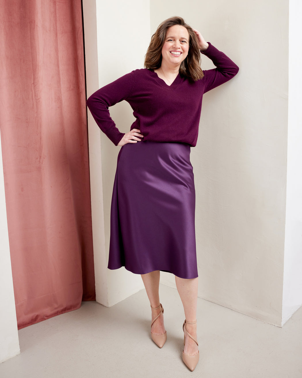 Woman wearing a purple sweater and skirt standing against a white wall with a pink curtain. This skirt is the Astell Bias Skirt sewing pattern from Cashmerette.