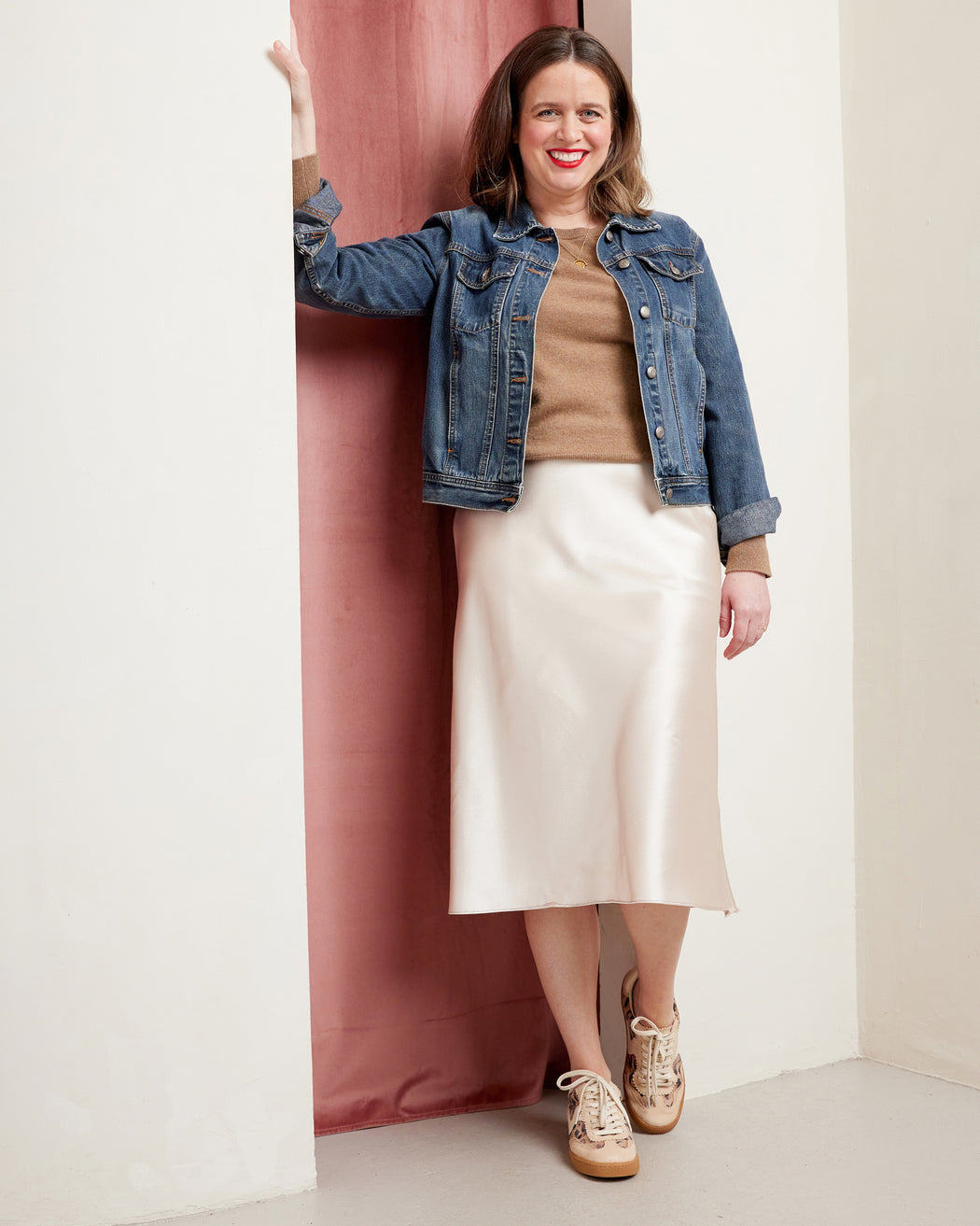 Woman wearing a denim jacket, beige sweater, and white skirt against a pink curtain. This skirt is the Astell Bias Skirt sewing pattern from Cashmerette.