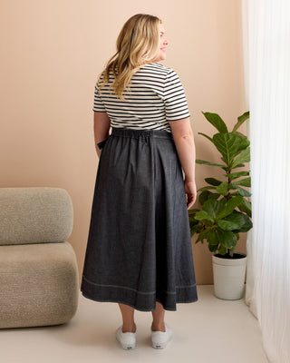 Woman wearing a striped shirt and dark skirt standing in a room with a plant and beige chair. This is the Osgood Skirt sewing pattern from Cashmerette.