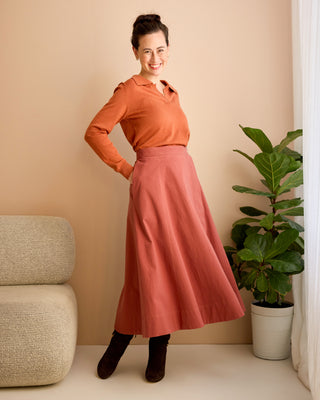 Woman in an orange blouse and pink skirt standing in a room with a beige wall and a plant. This is the Osgood Skirt sewing pattern from Cashmerette.