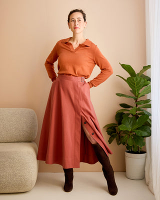 Woman wearing a rust-colored blouse and pink skirt standing in a room with a beige wall and a plant. This is the Osgood Skirt sewing pattern from Cashmerette.
