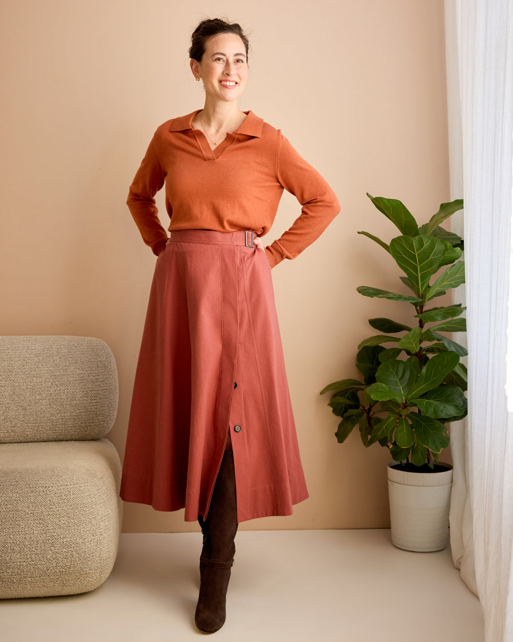 Woman wearing a rust-colored shirt and skirt standing in a room with a plant and beige sofa. This is the Osgood Skirt sewing pattern from Cashmerette.
