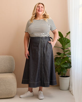 Woman wearing a striped shirt and dark denim skirt standing indoors with a plant and chair in the background. This is the Osgood Skirt sewing pattern from Cashmerette.