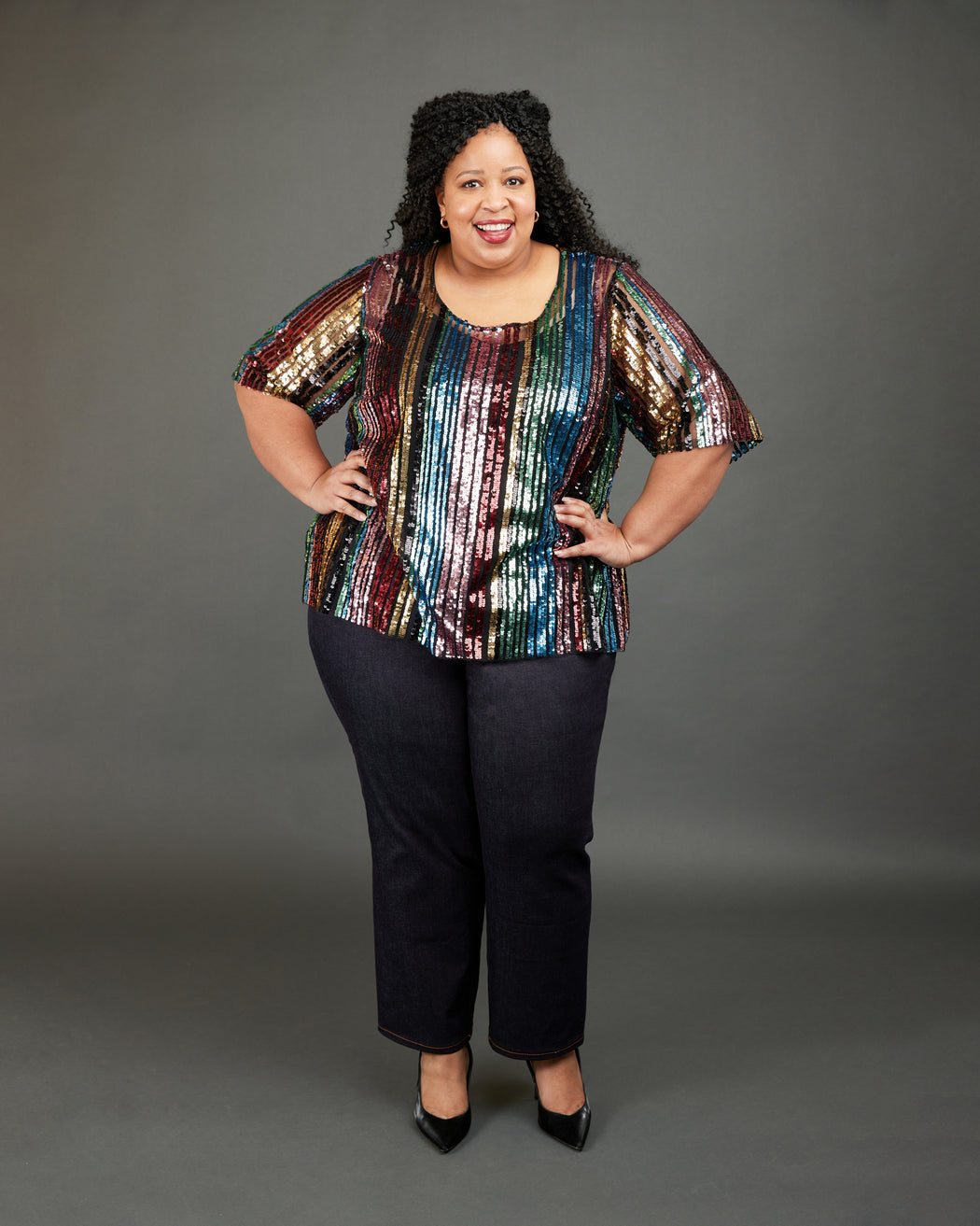 Woman wearing a colorful sequin top and black pants against a gray background. These jeans are the Ames Jeans sewing pattern from Cashmerette.