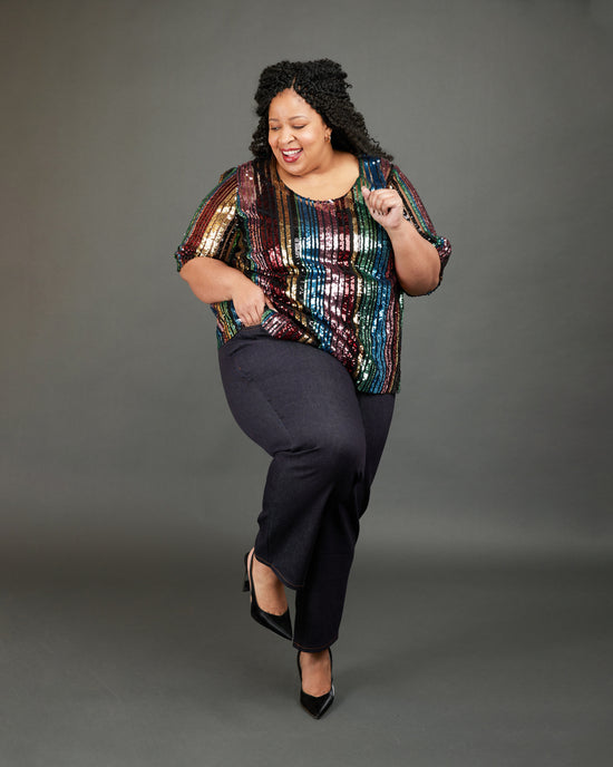 Person wearing a colorful sequin top and black pants on a gray background. These jeans are the Ames Jeans sewing pattern from Cashmerette.