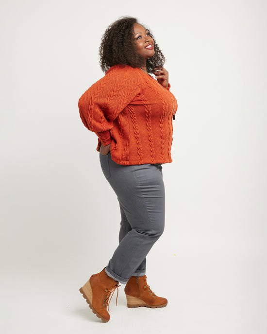 Woman wearing an orange cable knit sweater, gray pants, and brown boots on a white background. These jeans are the Ames Jeans sewing pattern from Cashmerette.