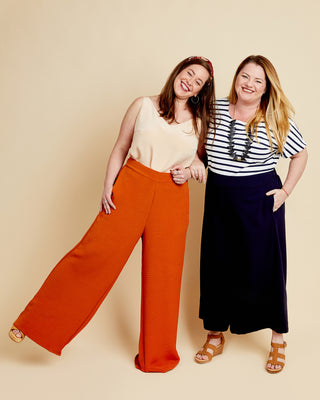 Two women standing side by side against a beige background, one wearing orange pants and a beige top, the other in a striped shirt and navy pants. These pants are the Calder Pants & Shorts sewing pattern from Cashmerette.