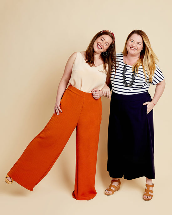 Two women standing side by side against a beige background, one wearing orange pants and a beige top, the other in a striped shirt and navy pants. These pants are the Calder Pants & Shorts sewing pattern from Cashmerette.