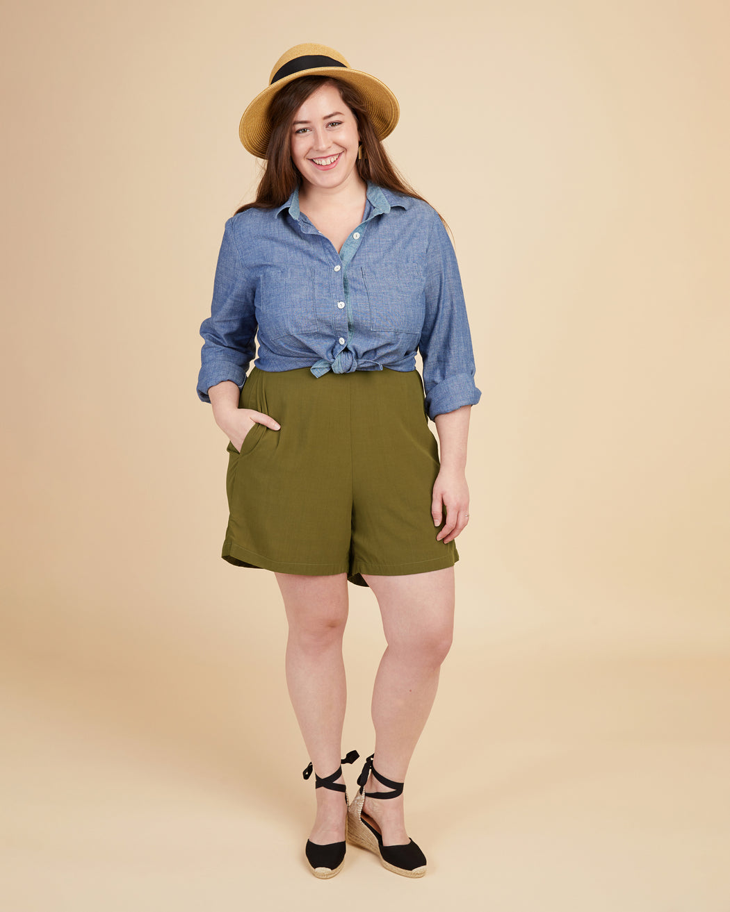 Woman wearing a blue shirt and green shorts with a beige hat on a beige background. These shorts are the Calder Pants & Shorts sewing pattern from Cashmerette.