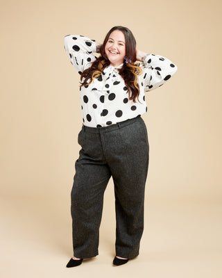 Person wearing a polka dot blouse and gray pants on a beige background. These pants are the Meriam Trousers sewing pattern from Cashmerette.