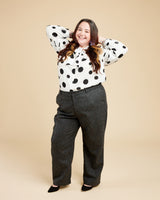 Person wearing a polka dot blouse and gray pants on a beige background. These pants are the Meriam Trousers sewing pattern from Cashmerette.