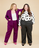 Two women posing together against a beige background, one in a purple suit and the other in a polka dot blouse and gray pants.. These pants are the Meriam Trousers sewing pattern from Cashmerette.
