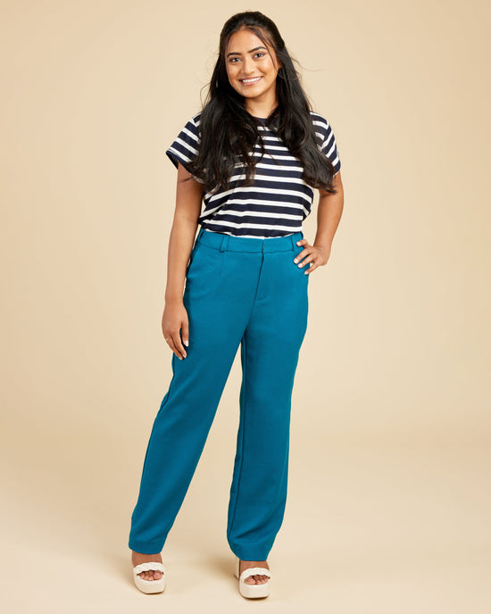 Woman wearing a black and white striped shirt with blue pants on a beige background. These pants are the Meriam Trousers sewing pattern from Cashmerette.