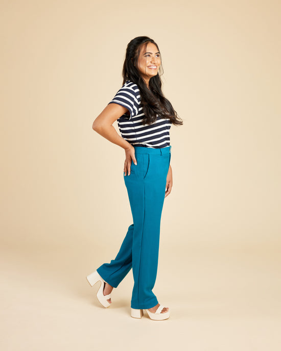 Woman wearing a black and white striped shirt and blue pants on a beige background. These pants are the Meriam Trousers sewing pattern from Cashmerette.