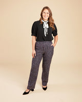 Woman wearing a black blouse with a white bow and patterned pants on a beige background. These pants are the Meriam Trousers sewing pattern from Cashmerette.