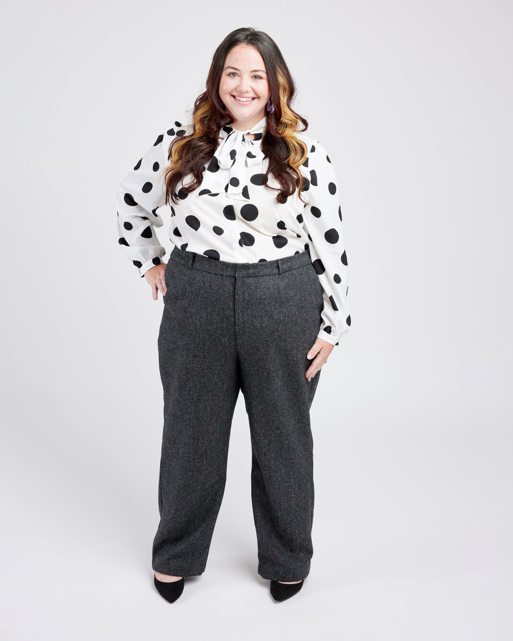Woman wearing a polka dot blouse and gray pants on a white background. These pants are the Meriam Trousers sewing pattern from Cashmerette.