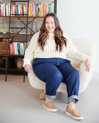 Woman sitting in a chair wearing a white sweater and blue pants, with a bookshelf in the background. These jeans are the Creston Jeans sewing pattern from Cashmerette.