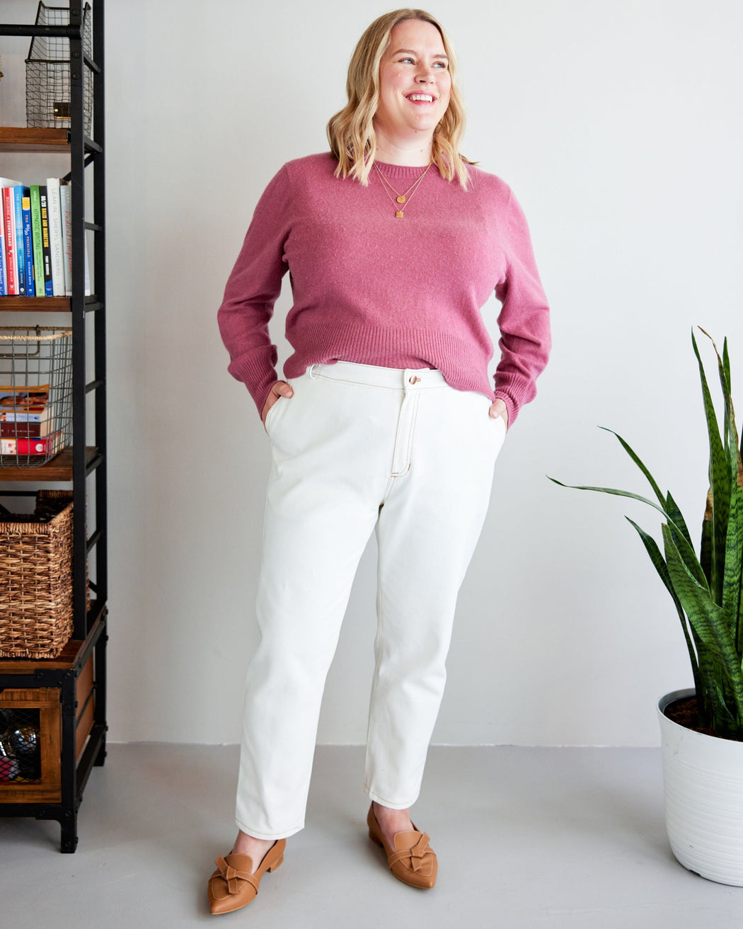 Woman wearing a pink sweater and white pants standing in a room with a bookshelf and plant. These jeans are the Creston Jeans sewing pattern from Cashmerette.