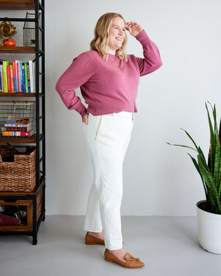 Woman wearing a pink sweater and white pants standing in a room with a bookshelf and plant. These jeans are the Creston Jeans sewing pattern from Cashmerette.