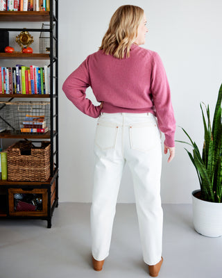 Woman wearing a pink sweater and white pants standing in a room with a bookshelf and plant. These jeans are the Creston Jeans sewing pattern from Cashmerette.