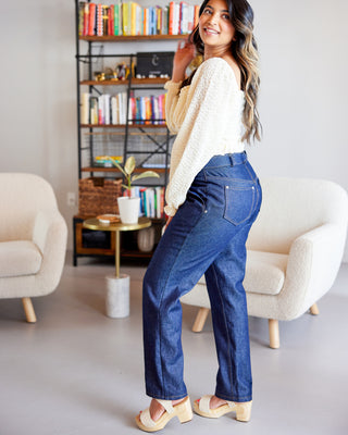 Woman in a cozy living room wearing blue jeans and a white sweater. These jeans are the Creston Jeans sewing pattern from Cashmerette.