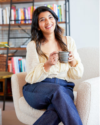 Woman sitting on a couch holding a mug in a cozy room with bookshelves. These jeans are the Creston Jeans sewing pattern from Cashmerette.