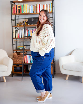 Woman in a white sweater and blue pants standing in a room with bookshelves and chairs. These jeans are the Creston Jeans sewing pattern from Cashmerette.