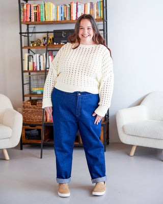 Woman wearing a cream sweater and blue pants standing in a room with bookshelves and chairs. These jeans are the Creston Jeans sewing pattern from Cashmerette.